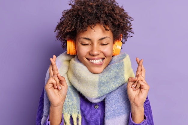 Zufriedenes afroamerikanisches Teenager-Mädchen hält die Augen geschlossen beißt Lippen steht abergläubisch kreuzt die Finger für viel Glück trägt Kopfhörer auf den Ohren Schal um den Hals