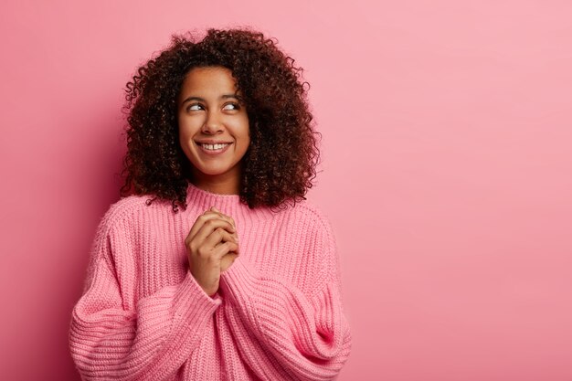 Zufriedenes dunkelhäutiges Mädchen mit lockigem Afro-Haar hält die Hände zusammen, konzentriert über dem Lächeln, trägt sanft gestrickte Pulloverträume über etwas Schönes, hat einen fröhlichen, nachdenklichen Ausdruck, der auf Pink isoliert ist