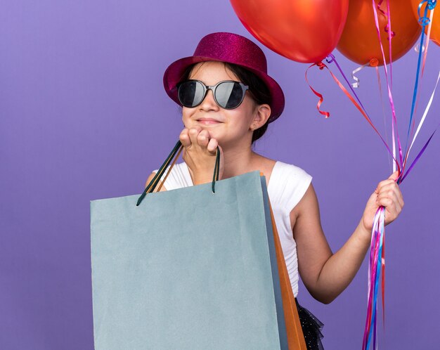 zufriedenes junges kaukasisches Mädchen in Sonnenbrille mit violettem Partyhut, das die Hand auf das Kinn legt und Heliumballons und Einkaufstüten isoliert auf lila Wand mit Kopierraum hält