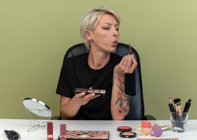 Zufriedenes junges schönes Mädchen sitzt am Tisch mit Make-up-Tools, die Make-up-Pinsel auf olivgrüner Wand halten und blasen