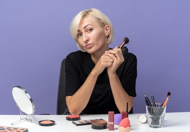 Zufriedenes junges schönes Mädchen sitzt am Tisch mit Make-up-Tools, die Pulverbürsten isoliert auf blauer Wand halten