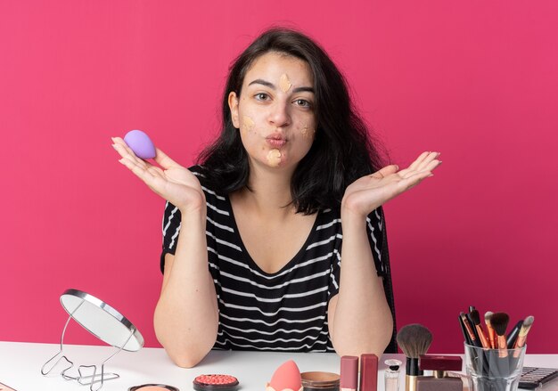 Zufriedenes junges schönes Mädchen sitzt am Tisch mit Make-up-Tools, die Ton-up-Creme auftragen, die einen Schwamm hält, der die Hände ausbreitet und die Kussgeste isoliert auf rosa Wand zeigt