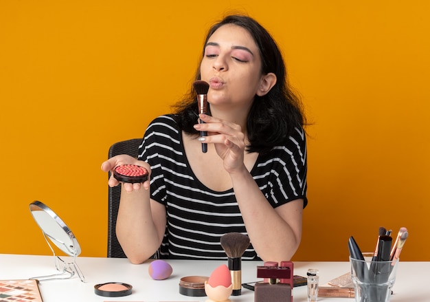 Zufriedenes junges, schönes Mädchen sitzt am Tisch mit Make-up-Werkzeugen, die Puderröte und Blasbürste in der Hand halten, isoliert auf oranger Wand