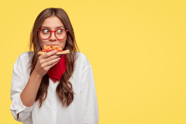 Zufriedenes kaukasisches Model isst köstliche Pizza drinnen, isst zu Mittag, trägt eine optische Brille, ein weißes Hemd und ein rotes Kopftuch, steht an der gelben Wand und bietet Platz für Ihren Slogan oder Text