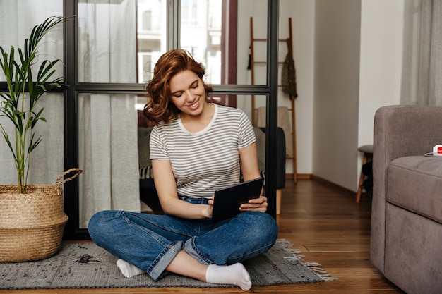 Zufriedenes Mädchen in Jeans und gestreiftem Oberteil sitzt auf dem Boden im Wohnzimmer und arbeitet im Laptop mit einem Lächeln.