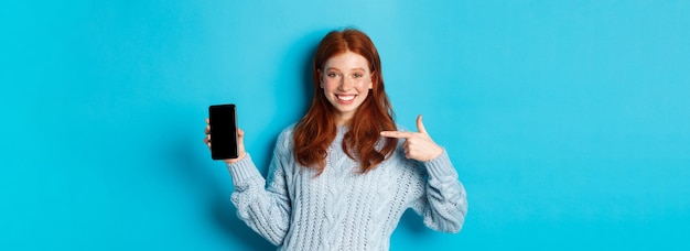 Kostenloses Foto zufriedenes rothaariges mädchen, das auf den telefonbildschirm zeigt, der smartphone-app oder online-promo zeigt und lächelt