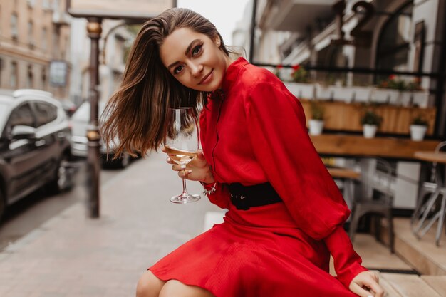 Zufriedenes, süßes Mädchen lächelt sanft. Rotes Kleid verleiht dem Outfit der Dame, die mit einem Glas Wein posiert, Helligkeit