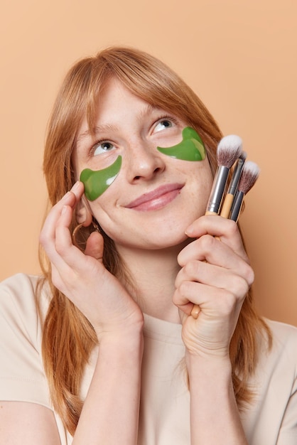 Zufriedenes, verträumtes Ingwermädchen berührt das Gesicht, trägt sanft grüne Hydrogelflecken unter den Augen auf, hält Kosmetikpinsel, die sich oben konzentrieren, und unterzieht sich Schönheitsverfahren zu Hause isoliert über braunem Hintergrund