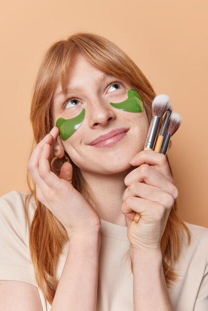 Zufriedenes, verträumtes Ingwermädchen berührt das Gesicht, trägt sanft grüne Hydrogelflecken unter den Augen auf, hält Kosmetikpinsel, die sich oben konzentrieren, und unterzieht sich Schönheitsverfahren zu Hause isoliert über braunem Hintergrund