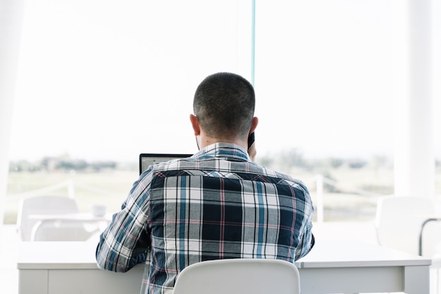 Zurück von einem Mann, der vor dem Laptop arbeitet und mit dem Mobile spricht