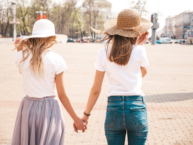 Zurück von zwei jungen schönen blonden lächelnden Hippie-Mädchen in der weißen T-Shirt Kleidung und im Hut des modischen Sommers. . Paar einander die Hände halten