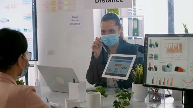 Zurückaufnahme von Geschäftsfrauen mit medizinischer Gesichtsmaske, die bei der Präsentation des Managements mit Tablet-Computer zusammenarbeiten, während sie im Firmenbüro sitzen. Team respektiert soziale Distanz