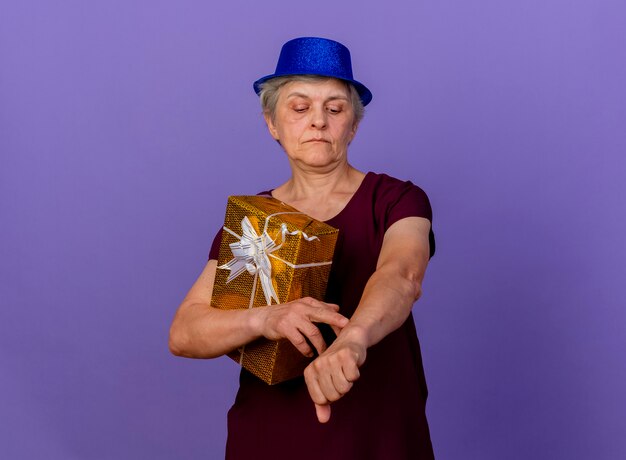 Zuversichtlich ältere Frau, die Parteihut trägt, hält Geschenkbox, die Arm lokalisiert auf lila Wand mit Kopienraum betrachtet