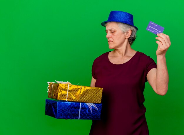 Zuversichtlich ältere Frau, die Partyhut trägt, hält Geschenkboxen und Kreditkarte auf Grün