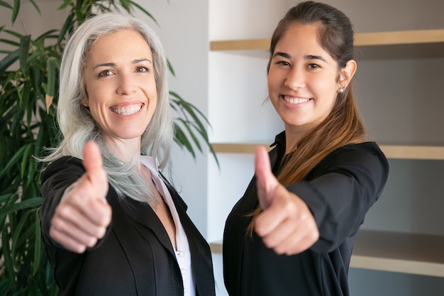 Zuversichtlich Büro Arbeitgeber Daumen hoch und lächelnd. Zwei glückliche professionelle Geschäftsfrauen, die zusammen stehen und im Besprechungsraum posieren. Konzept für Teamarbeit, Geschäft und Zusammenarbeit