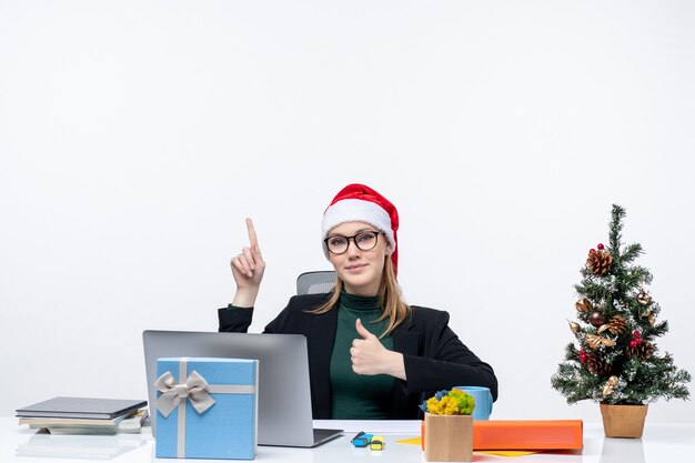 Zuversichtlich Geschäftsfrau mit Weihnachtsmannhut, der an einem Tisch mit einem Weihnachtsbaum und einem Geschenk darauf sitzt, macht ok Geste und zeigt etwas im Büro auf weißem Hintergrund