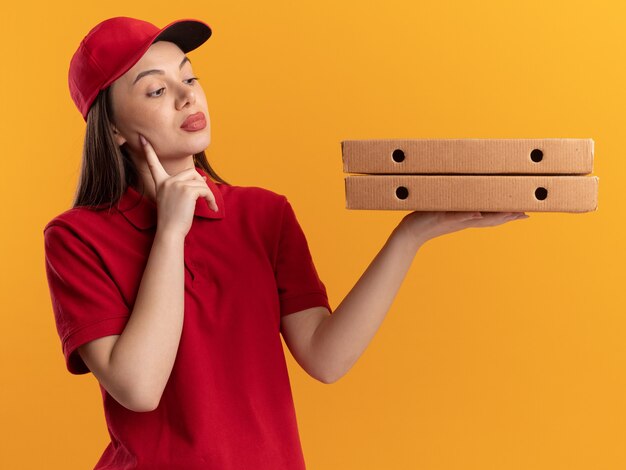 Zuversichtlich hübsche Lieferfrau in Uniform legt Finger auf Gesicht, das Pizzaschachteln auf Orange hält und betrachtet