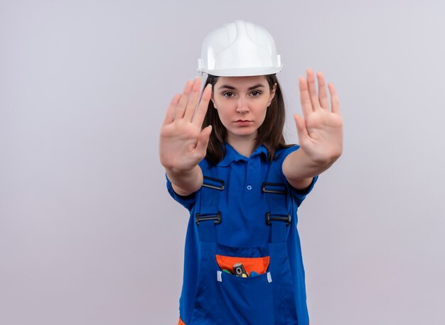 Zuversichtlich junge Baumeisterin mit weißem Schutzhelm und blauen Uniformgesten halten mit beiden Händen auf lokalisiertem weißem Hintergrund mit Kopienraum an