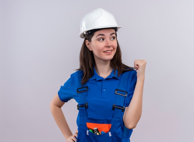 Zuversichtlich junge Baumeisterin mit weißem Schutzhelm und blauer Uniform erhebt Faust auf lokalisiertem weißem Hintergrund mit Kopienraum