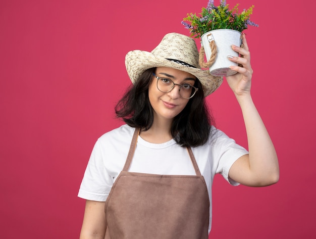 Zuversichtlich junge brünette Gärtnerin in optischen Gläsern und in Uniform, die Gartenhut trägt, hält Blumentopf über Kopf lokalisiert auf rosa Wand