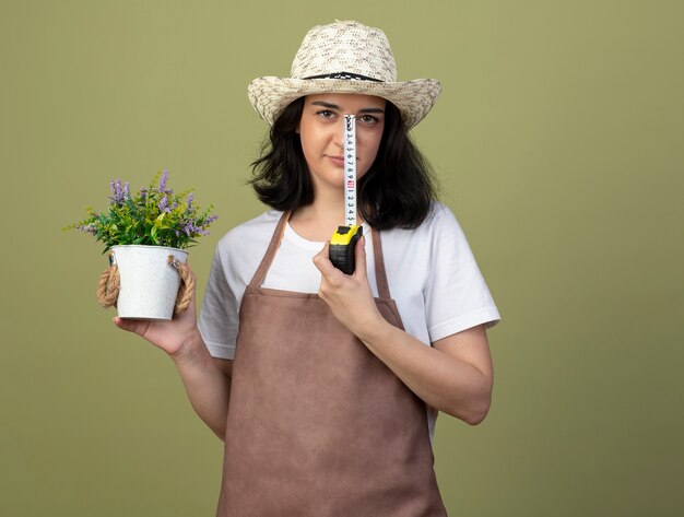 Zuversichtlich junge brünette weibliche Gärtnerin in Uniform mit Gartenhut hält Blumentopf und Maßband isoliert auf olivgrüner Wand