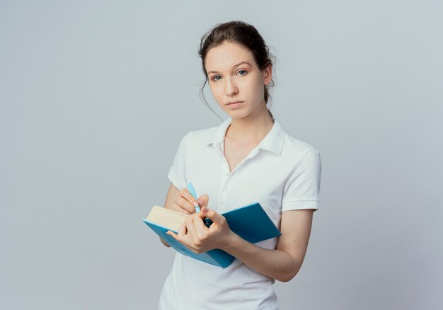 Zuversichtlich junge hübsche Studentin, die offenes Buch und Stift hält und Kamera lokalisiert auf weißem Hintergrund mit Kopienraum betrachtet