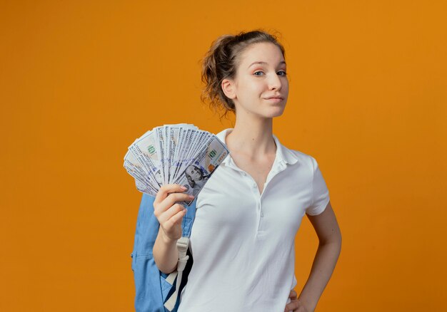 Zuversichtlich junge hübsche Studentin, die Rückentasche hält Geld hält Hand auf Taille lokalisiert auf orange Hintergrund mit Kopienraum