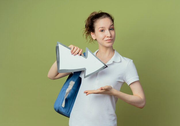 Zuversichtlich junge hübsche Studentin, die Rückentasche hält Pfeilmarkierung, die zur Seite zeigt und mit der Hand darauf zeigt, lokalisiert auf olivgrünem Hintergrund