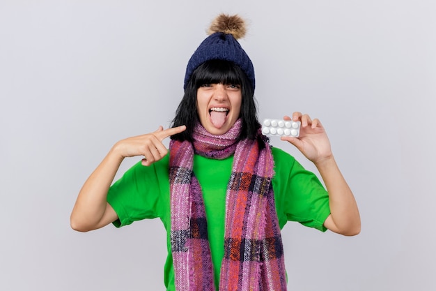 Zuversichtlich junge kranke Frau, die Wintermütze und Schal trägt, die Packung der Tabletten zeigt, die auf sie schauen, die Front zeigt Zunge lokalisiert auf weißer Wand