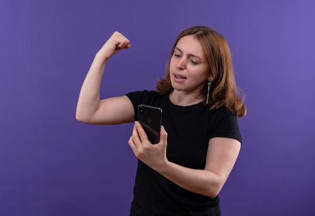 Zuversichtlich junge lässige Frau, die Handy hält und starke Geste tut, die Handy auf lokalisiertem lila Raum mit Kopienraum betrachtet