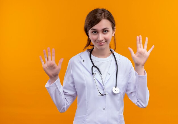 Zuversichtlich junge Ärztin, die medizinische Robe und Stethoskop trägt, die leere Hände auf lokalisiertem orange Raum zeigen