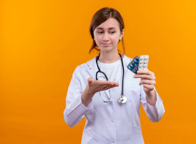 Zuversichtlich junge Ärztin, die medizinische Robe und Stethoskop trägt, die medizinische Drogen halten und mit der Hand auf sie auf isoliertem orange Raum mit Kopienraum zeigen