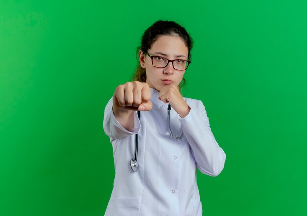 Zuversichtlich junge Ärztin, die medizinische Robe und Stethoskop und Brille trägt, die Boxgeste tut, lokalisiert auf grüner Wand mit Kopienraum