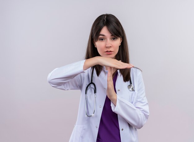 Zuversichtlich junge Ärztin im medizinischen Gewand mit Stethoskop gestikuliert Zeitüberschreitung auf lokalisiertem weißem Hintergrund mit Kopienraum