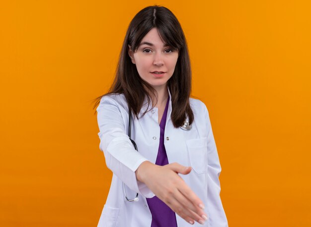 Zuversichtlich junge Ärztin im medizinischen Gewand mit Stethoskop hält Hand auf lokalisiertem orange Hintergrund mit Kopienraum aus