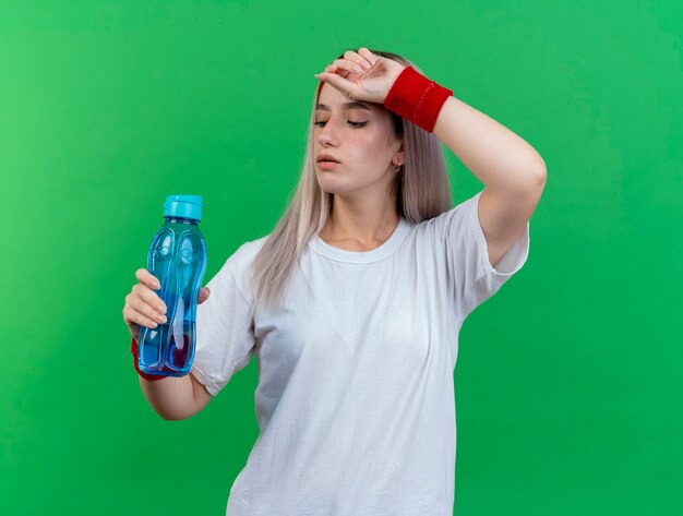 Zuversichtlich junge sportliche Frau mit Zahnspangen, die Stirnband und Armbänder tragen, legt Hand auf Stirn und schaut auf Wasserflasche isoliert auf grüner Wand