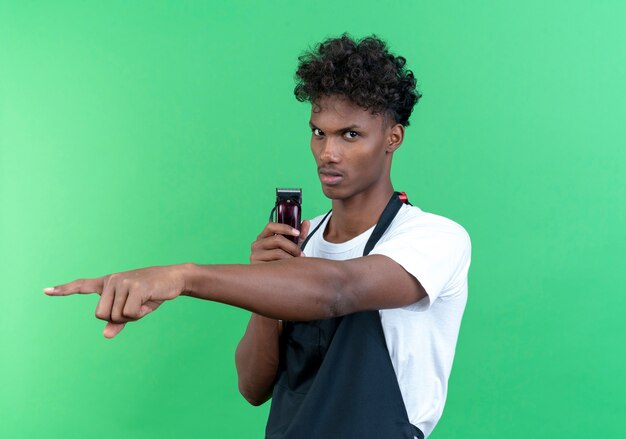 Zuversichtlich junger afroamerikanischer männlicher Friseur, der Uniform hält Haarschneidemaschinen und Punkte seitlich lokalisiert auf grünem Hintergrund