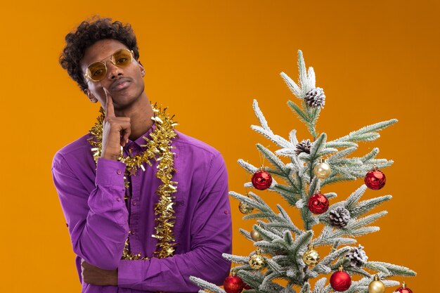 Zuversichtlich junger afroamerikanischer Mann, der eine Brille mit Lametta-Girlande um den Hals trägt, die nahe verziertem Weihnachtsbaum berührt Gesicht berührt auf orange Wand steht