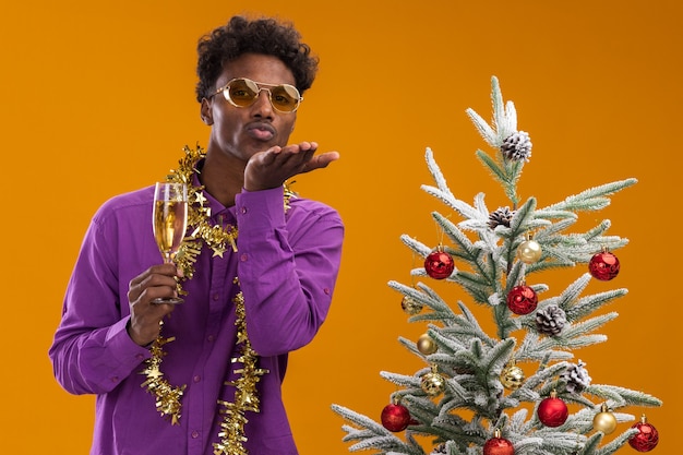 Zuversichtlich junger afroamerikanischer Mann, der eine Brille mit Lametta-Girlande um den Hals trägt, die nahe verziertem Weihnachtsbaum steht, der Glas Champagner hält