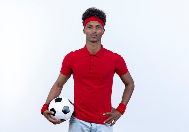 Kostenloses Foto zuversichtlich junger afroamerikanischer sportlicher mann, der haarband und armband hält ball hält und hand auf hüfte lokalisiert auf weißem hintergrund setzt