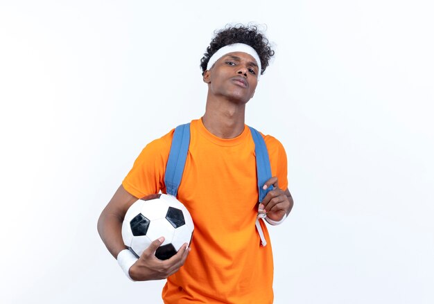 Zuversichtlich junger afroamerikanischer sportlicher Mann, der Stirnband und Armband und Rückentasche hält Ball lokalisiert auf weißem Hintergrund trägt