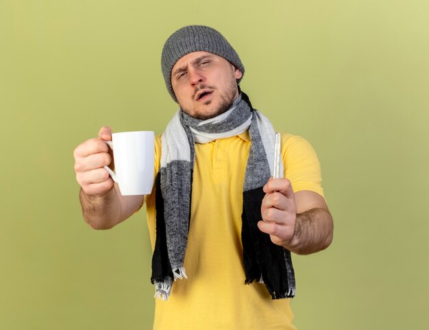 Zuversichtlich junger blonder kranker slawischer Mann, der Wintermütze und Schal trägt, hält Tasse und Thermometer lokalisiert auf olivgrüner Wand mit Kopienraum