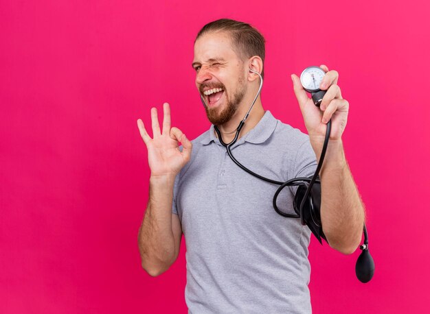 Zuversichtlich junger gutaussehender slawischer kranker Mann, der Stethoskop trägt, das seinen Druck mit Blutdruckmessgerät misst, das vorne zwinkert und ok singt auf rosa Wand mit Kopienraum