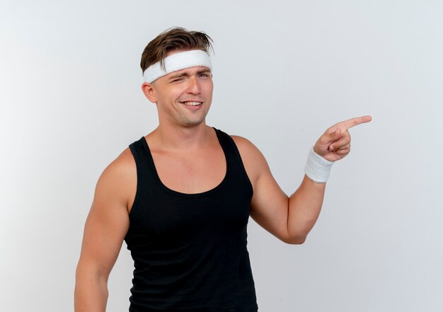 Zuversichtlich junger gutaussehender sportlicher Mann, der Stirnband und Armbänder lächelt und zur Seite zeigt, die auf Weiß lokalisiert wird