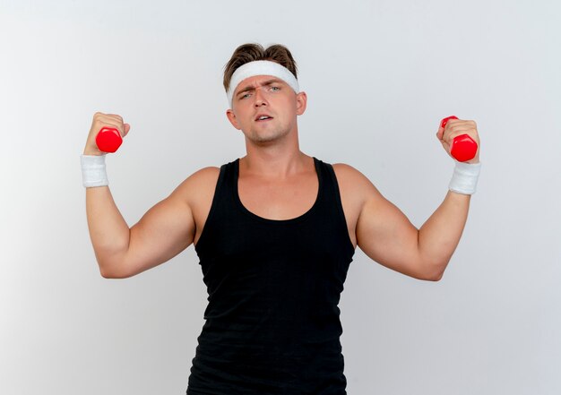 Kostenloses Foto zuversichtlich junger gutaussehender sportlicher mann, der stirnband und armbänder trägt, die hanteln lokalisiert auf weiß anheben