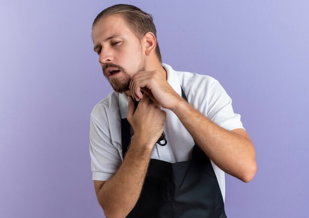 Zuversichtlich junger hübscher Friseur, der Uniform hält, die Haarschneidemaschinen hält, die seinen eigenen Bart schneiden und sein Kinn berühren, das Seite betrachtet, die auf Purpur mit Kopienraum isoliert ist