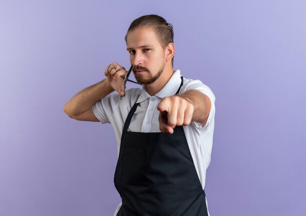 Zuversichtlich junger hübscher Friseur, der Uniform hält, die Schere nahe Gesicht hält und lokalisiert auf Purpur mit Kopienraum zeigt