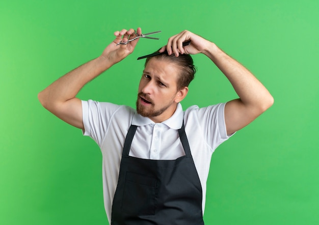 Zuversichtlich junger hübscher Friseur, der Uniform trägt, die sein Haar kämmt und Schere hält, die auf Grün isoliert wird