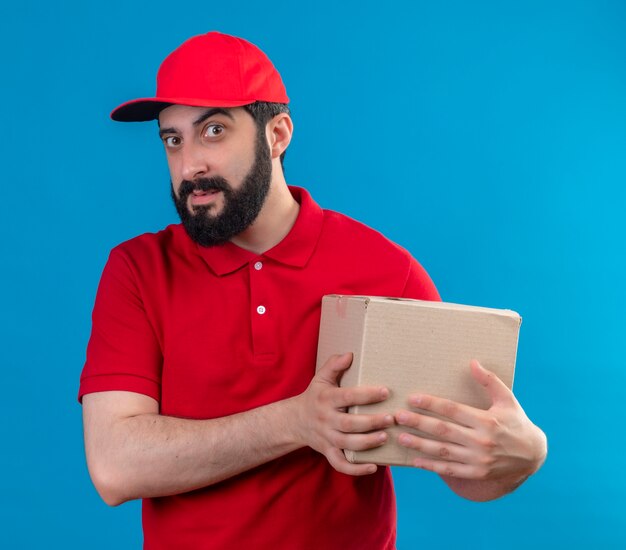 Zuversichtlich junger hübscher kaukasischer Lieferbote, der rote Uniform und Kappe hält Kartonbox auf Blau isoliert trägt