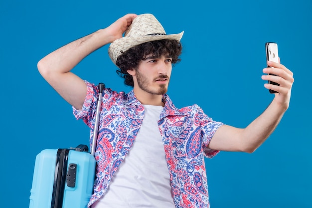Zuversichtlich junger hübscher lockiger reisender Mann, der Hut nimmt, der selfie Hand auf Hut mit Koffer auf isoliertem blauem Raum setzt